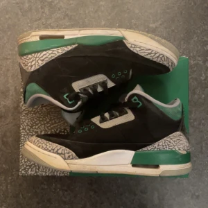 Air Jordan 3 Retro - Snygga Air Jordan 3 Retro sneakers i svart med detaljer i grönt och silver. Skorna har en klassisk design med snörning och en unik mönstrad sula. Perfekta för dig som vill ha en stilren och sportig look.