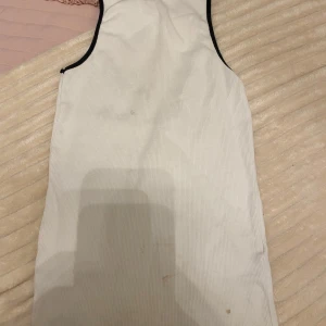 Vit ärmlös klänning - Säljer en stilren vit ärmlös tröja  med svarta kanter. Perfekt för en minimalistisk look. Tröjan har en enkel och elegant design som passar till många tillfällen. Men den har små fläck