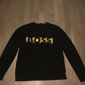 Svart tröja från BOSS med gulddetaljer - Snygg svart tröja från BOSS med ett iögonfallande guldtryck på bröstet. Tröjan har långa ärmar och en rund halsringning. Perfekt för en stilren look. Storlek S. Ny pris 1849 mitt pris 1099