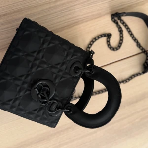 ”INTE DEN RIKTIGA ”Svart handväska med kedja - Snygg svart handväska med quiltad design och kedjedetaljer. Väskan har en elegant form och ett rött foder inuti. Perfekt för att ge en stilren touch till din outfit.