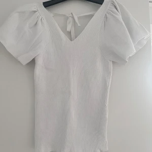 Vit ribbad topp från Only - Säljer en stilren vit ribbad topp från Only med korta puffärmar och v-ringning. Toppen har en knytning i ryggen som ger en fin detalj. Sitter väldigt bra❤️❤️hör av dig om du har några frågor!