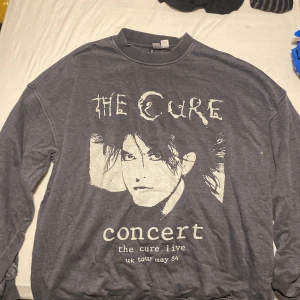 Grå sweatshirt med The Cure-tryck från Divided - Säljer en grå sweatshirt från Divided (H&M) med ett coolt The Cure-tryck på framsidan. Tröjan har långa ärmar och en rund halsringning. Perfekt för fans av bandet eller för en avslappnad stil. 🖤 (lOversized och stor dock!) 