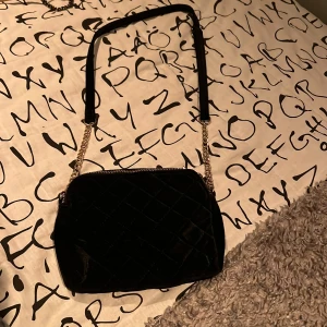 Svart quiltad axelväska med guldkedjedetaljer - Snygg svart axelväska i quiltat material med guldfärgade kedjedetaljer. Väskan har två dragkedjor upptill och en justerbar axelrem. Använd 1 gång. Används inte för att jag är en silverperson. 