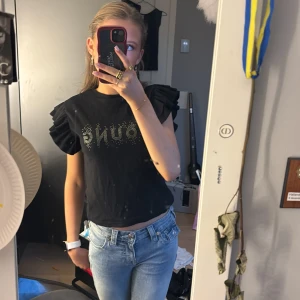 Svart t-shirt med volangärmar - Snygg svart t-shirt från Gina med texten 'YOUNG' i glittriga stenar på framsidan. Den har korta volangärmar som ger en extra touch. Perfekt för en trendig look!