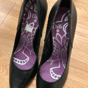 Svarta pumps från Chilli - Snygga svarta pumps från Chilli med spetsig tå och hög klack. Skorna har en lila innersula med ett dekorativt mönster. Perfekta för en elegant look.