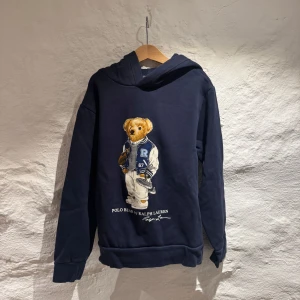 Mörkblå hoodie från Ralph Lauren - Säljer en snygg mörkblå hoodie från Ralph Lauren med en ikonisk björn på framsidan. Tröjan har långa ärmar och en bekväm huva. Den e i bra skick och ädast använd varsamt, pris kan diskuteras och var nu inte rädd att höra av dig vid några funderingar!🤩🍸🍾🐎