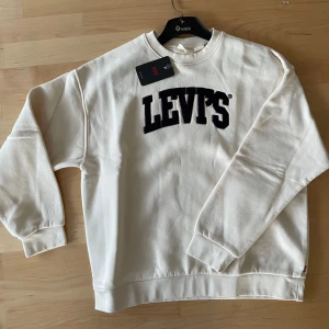 Vit sweatshirt från Levi's S oanvänd med prislapp  - Säljer en stilren vit sweatshirt från Levi's med deras ikoniska logga i svart på bröstet. Tröjan har långa ärmar och en klassisk rund halsringning. Perfekt för en avslappnad stil.
