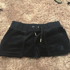 Svarta sammetsshorts med fickor fina till sommaren - Snygga svarta sammetsshorts från Juicy Couture med elastisk midja och snörning. Shortsen har praktiska fickor både fram och bak, med knappar på bakfickorna. Perfekta för en avslappnad stil.