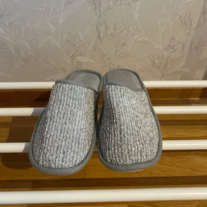 Grå tofflor i syntetmaterial - Mysiga grå tofflor i syntetmaterial med en ribbad ovansida. Perfekta för att hålla fötterna varma inomhus. Sulan har ett mönster för bättre grepp.