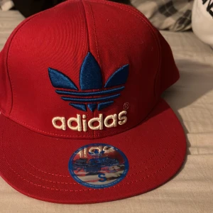 Röd keps från Adidas - Snygg röd keps från Adidas med blått och vitt broderat logotyp framtill. Kepsen har en klassisk design med böjd skärm och justerbar passform. Perfekt för en sportig look!