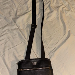 Svart axelväska från Emporio Armani - Snygg svart axelväska från Emporio Armani i skinn med justerbar axelrem. Väskan har en dragkedja upptill och en extra ficka med dragkedja på framsidan. Perfekt för att bära dina nödvändigheter med stil.