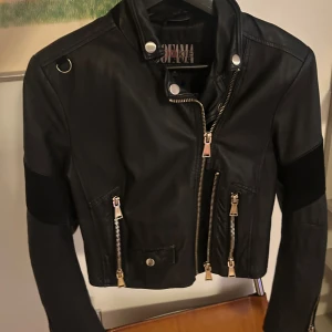 Kort skinnjacka från Jofama - Snygg svart skinnjacka från Jofama x Rookie med coola dragkedjedetaljer och broderade rosor på insidan. Jackan har en croppad passform och långa ärmar. Perfekt för en edgy look!