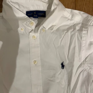 Vit skjorta från Ralph Lauren - Klassisk vit skjorta från Ralph Lauren med broderad logotyp på bröstet. Skjortan har långa ärmar och knappar framtill. Perfekt för en stilren look.