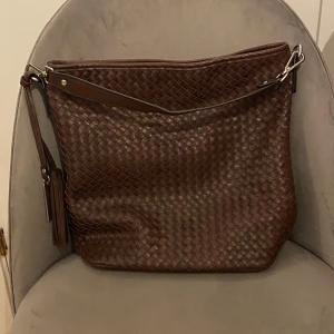 Brun shoppingväska Suri Frey!  - Snygg brun axelväska i skinn med flätad design på framsidan och slät baksida. Väskan har en praktisk dragkedja och en dekorativ tofs. Insidan är fodrad med ett mönstrat tyg och har flera fack för enkel organisering.Marley är namnet på väskan Höjd 34 cm  2 fickor inuti väska varav 1 med dragkedja  1 ficka med dragkedja på utsidan  Jättefin väska! Mycket bra skick!  