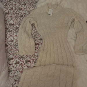 Vit stickad klänning - Mysig beige stickad klänning med kabelstickat mönster. Klänningen är figurnära och ger en elegant siluett. Ny med prislapp, i strl XS