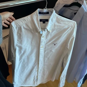 Vit skjorta från GANT - Säljer en klassisk vit skjorta från GANT i storlek S. Skjortan har långa ärmar och knappar framtill samt en broderad logotyp på bröstet. Perfekt för en stilren look.