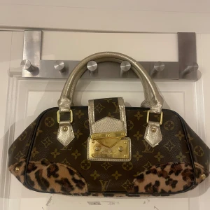 Brun handväska med leoparddetaljer - Snygg brun handväska med  leoparddetaljer. Väskan har guldfärgade metalldetaljer och ett handtag i glittrigt material. Insidan är fodrad med leopardmönster och har en dragkedja för extra säkerhet.