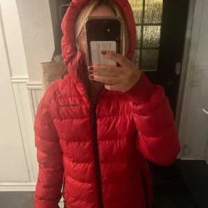 Röd peak performance jacka med huva - Snygg röd dunjacka från Peak Performance med huva och dragkedja. Perfekt för kyliga dagar. Jackan har en pufferdesign och långa ärmar.