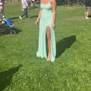 Mintgrön långklänning med öppen rygg - Elegant mintgrön långklänning med smala axelband och en djärv slits. Klänningen har en öppen rygg med snörning och glittrigt mönster som fångar ljuset vackert. Köpt 2024 på dressroomlinné Göteborg. Använd 1 gång. Nypris 6000