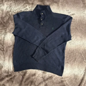 Polo Ralph Lauren - Säljer en snygg mörkblå tröja från Polo Ralph Lauren i storlek L. Tröjan är tillverkad i 100% bomull och har en ribbad krage med knappar. Perfekt för kyligare dagar med sin stilrena design och långa ärmar.