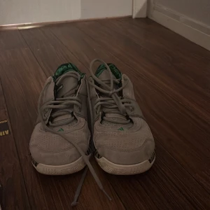 Adidas Gil Zero - Säljer ett par gråa Adidas med gröna detaljer och zebramönster på hälen. Skorna har snörning och en unik sula med gröna cirkulära mönster. Perfekta för en sportig look.