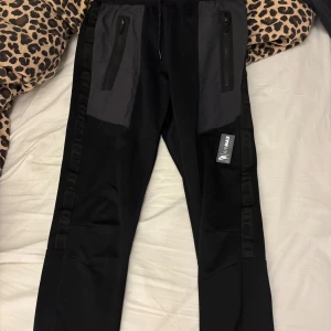Svarta träningsbyxor från Armax - Snygga svarta träningsbyxor från Armax med dragkedjor på fickorna och en bekväm passform. Perfekta för gymmet eller löprundan. Byxorna har en elastisk midja och är gjorda i ett slitstarkt material.