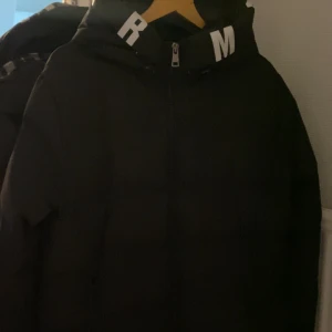 Svart dunjacka med textdetaljer - Snygg svart dunjacka med textdetaljer på kragen. Perfekt för kalla dagar med sin varma och vadderade design. Jackan har en dragkedja framtill och en hög krage för extra skydd mot kyla.