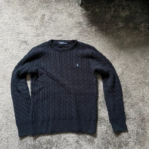 Svart kabelstickad tröja från Ralph Lauren - Snygg svart kabelstickad tröja från Ralph Lauren med rund hals och det klassiska logotypbroderiet på bröstet. Perfekt för kyligare dagar och ger en stilren look. Storlek S-M