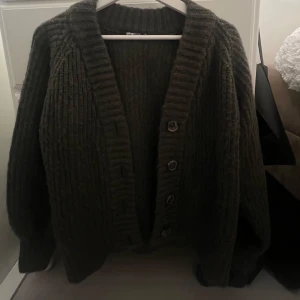 Mörkgrön stickad kofta med knappar - Säljer en mysig mörkgrön stickad kofta med stora knappar framtill. Koftan har en ribbad struktur och långa ärmar, perfekt för kyliga dagar. Den har en klassisk design som passar till många olika stilar.