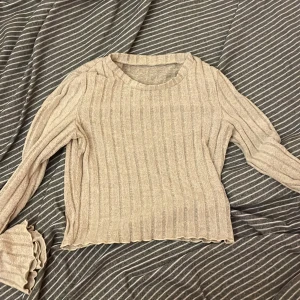Beige ribbad tröja - Säljer en beige ribbad tröja med långa ärmar. Tröjan har en rund halsringning och är i ett mjukt material som passar perfekt för kyligare dagar. Den har en stilren design med vågiga kanter vid ärmslut och nederkant.