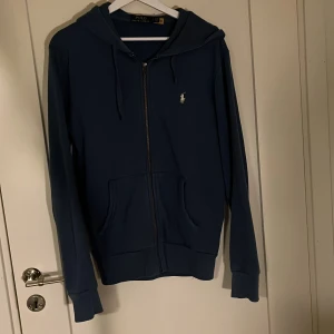 Blå hoodie från Polo Ralph Lauren - Säljer en stilren blå hoodie från Polo Ralph Lauren med dragkedja och huva. Tröjan har en liten logga på bröstet och praktiska fickor framtill. Perfekt för en avslappnad look.