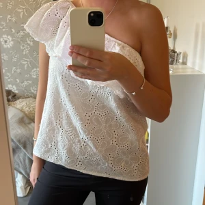 Vit oneshoulder blus med spetsdetaljer - Snygg vit oneshoulder blus med vacker spets och volangdetalj vid axeln. Perfekt för sommarens alla tillfällen. Blusen har en luftig passform och är tillverkad i ett lätt material.