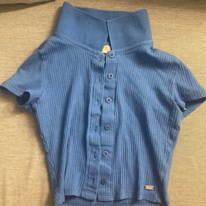 Blå ribbad topp från Pull&Bear - Säljer en blå ribbad topp från Pull&Bear med korta ärmar och knappar framtill. Den har en unik krage som ger en cool look. Perfekt för en avslappnad stil.