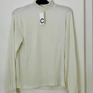 Vit långärmad blus från Cubus. - Off-white blus med långa ärmar och hög hals. Smockdetaljer vid hals och ärmslut. Ny med prislapp.