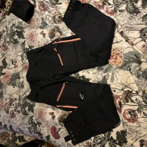 Svarta Vandringsbyxor  - Snygga svarta byxor från RevolutionRace med orange dragkedjor. Perfekta för utomhusaktiviteter med flera praktiska fickor och en bekväm passform. Designade i Sverige och tillverkade i Myanmar.