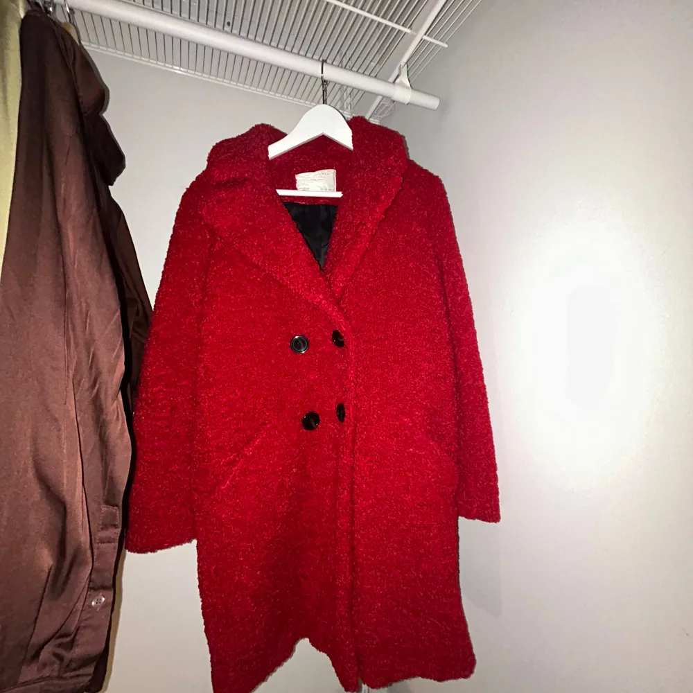 Säljer en mysig röd teddyjacka med stora svarta knappar. Jackan har en avslappnad passform och är perfekt för kyliga dagar. Den har långa ärmar och en skön krage som håller dig varm. Perfekt för att ge lite färg till din vintergarderob!. Takit.