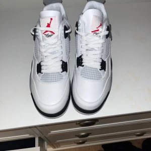 Vita Nike Air Jordans 4  - Snygga vita Nike Air Jordans med grå och svarta detaljer. Skorna har en klassisk design med snörning och den ikoniska Jumpman-loggan på plösen. Perfekta för sneaker-entusiaster som vill ha en stilren look.