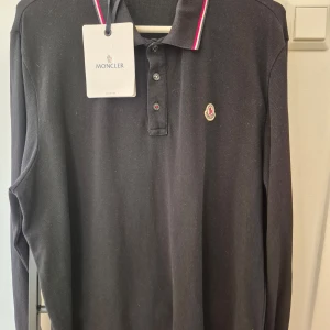 Svart långärmad pikétröja från Moncler - Snygg svart långärmad pikétröja från Moncler med klassisk krage och knappar. Tröjan har Monclers logotyp broderad på bröstet och en stilren design med röd och vit rand på kragen. NFC scan