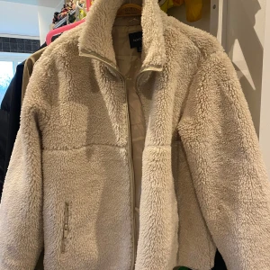 Beige teddyjacka från Boohoo - Mysig beige teddyjacka från Boohoo med dragkedja framtill. Perfekt för kyliga dagar med sin fluffiga och varma design. Jackan har långa ärmar och en hög krage för extra värme.