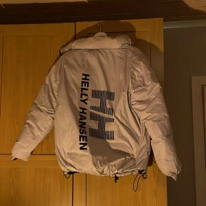Beige och svart pufferjacka från Helly Hansen - Säljer en vändbar pufferjacka från Helly Hansen i vit som har reflex i mörkret och svart. Jackan har en dragkedja framtill och justerbara snören i nederkant. Perfekt för kyliga dagar med sin vadderade design och långa ärmar.