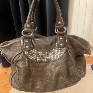 Brun skinnväska från Liebeskind - Snygg brun skinnväska från Liebeskind Berlin med nitar som detalj. Väskan har två robusta handtag och en rymlig design, perfekt för att bära med sig allt man behöver. Inuti väskan finns ett sidofack med drag kedja, två andra sidofack utan dragkedja samt ett större fack i mitten av väskan med dragkedja. Väskan har inget tillhörande längre band. 