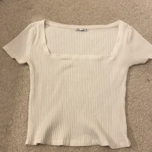 Vit ribbad topp från Zara - Säljer en stilren vit ribbad topp från Zara. Toppen har en fyrkantig halsringning och korta ärmar, perfekt för en avslappnad look. Tillverkad i ett mjukt och bekvämt material.