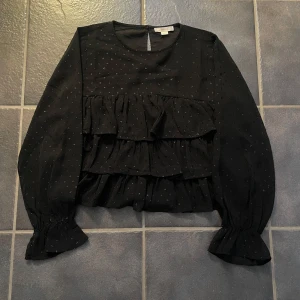 Svart blus med volanger från Gina Tricot - Snygg svart blus från Gina Tricot med volanger och diskreta prickar. Blusen har långa ärmar och en rund halsringning. Perfekt för en stilren look.