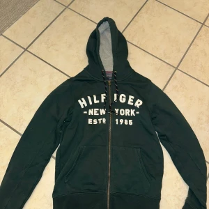 Grön hoodie från Tommy Hilfiger - Säljer en grön hoodie från Tommy Hilfiger med texten 'Hilfiger New York Estd. 1985' på framsidan. Den har en dragkedja och en klassisk känguruficka.