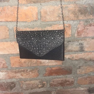 Svart glittrig kuvertväska med kedja - Snygg svart kuvertväska med glittriga detaljer och en elegant kedja som axelrem. Perfekt för en kväll ute eller speciella tillfällen. Väskan har en praktisk knappstängning och ett rymligt inre fack.