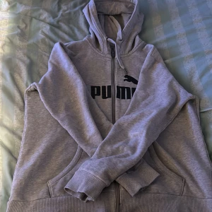 Grå hoodie från Puma - Säljer en grå hoodie från Puma med dragkedja och svart logga på bröstet. Tröjan har en bekväm passform med långa ärmar och en praktisk huva. Perfekt för en avslappnad stil.