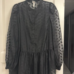 Svart prickig klänning - Elegant svart klänning med prickigt mönster och långa ärmar. Klänningen har knappar framtill och en luftig passform. Perfekt för höst och vinter.