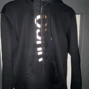 Svart hoodie från Hugo - Snygg svart hoodie från Hugo med dragkedja och stort tryck i silver och guld på framsidan. Perfekt för en stilren och avslappnad look. Den har en justerbar huva med snören och en praktisk ficka framtill.