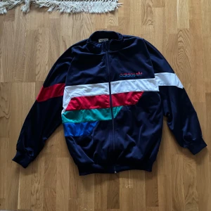 Flerfärgad träningsjacka från Adidas - Snygg träningsjacka från Adidas i blått med färgglada detaljer i rött, vitt, grönt och blått. Jackan har dragkedja och långärmade ärmar, perfekt för en sportig look.