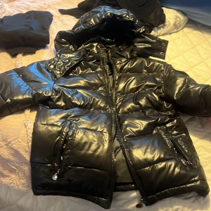Svart pufferjacka moncler  - Snygg svart pufferjacka från Moncler med glansig yta och praktisk huva. Jackan har dragkedja och flera fickor med dragkedjor för säker förvaring. Perfekt för kyliga dagar. Köpte den för mindre än en vecka sen men den passar inte mig 